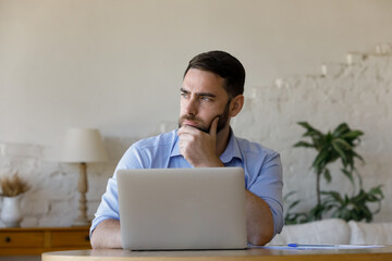 Pensive young businessman entrepreneur worker looking in distance stuck with difficult task, considering problem solution or creating project online, sitting at table with computer at home office.