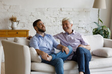 Happy Relaxed Young Man Laughing, Enjoying Pleasant Conversation With Older Retired Father, Resting Together On Cozy Couch. Joyful Carefree Two Generations Family Discussing Life News At Home.