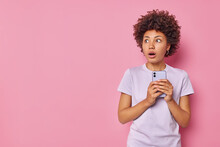 Horizontal Shot Of Scared Stunned Woman Holds Mobile Phone Sees Something Scary Wears Casual T Shirt Isolated Over Pink Background With Copy Space For Your Advertising Content Hears Bad News