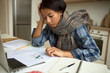© Anatoliy Karlyuk - Close-up of pretty sick young dark-skinned woman in scarf around neck sitting at kitchen in home office in front of laptop, studying papers, documents, self-isolated from corporate office life