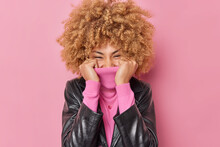 Positive Woman Hides Face With Collar Of Jumper Laughs Happily Wears Black Leather Jacket Feels Glad Isolated Over Pink Background. Joyful Female Model In Fashionable Clothes. Emotions Concept