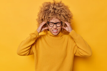 Wall Mural - Photo of displeased woman keeps hands on temples suffers from migraine clenches teeth to reveal pain cannot concentrate in noisy atmosphere dressed in casual jumper isolated over yellow wall