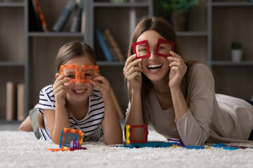 Wall Mural - Having fun. Funny portrait of happy young single mother schoolgirl daughter lying on floor looking at camera through magnet constructor pieces. Loving foster mom tween adopted kid play cute binoculars