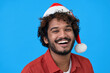 © insta_photos - Happy joyful young indian man smiling looking at camera wearing red santa hat advertising Christmas time shopping, xmas celebration party isolated on blue background. Headshot portrait.