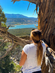 Wall Mural - Vertical shot of a female leaning on a tree looking at the blue lake and dense trees