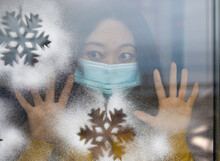 Sad Asian Girl With Medical Mask Wearing Yellow Sweater Looking Through Window In Christmas By The Coronavirus Pandemic