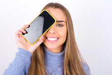 Young Caucasian Girl Wearing Blue Turtleneck Over White Background Holding Modern Smartphone Covering One Eye While Smiling