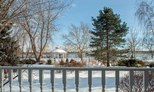 Winter Scene With Snow.  A Gazebo Sits In The Front Yard With Trees Around And A River In The Background.  In The Foreground Is A Wood Rail From A Verandah.
