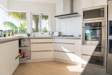 New Domestic White Interior Of Kitchen In Modern Home. With Sunlight Streaming In And View Through Windows.