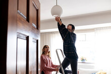 Young Woman Adjusting Pendant Light With Friend At Home