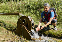 Elderly Adventurer With A Backpack Kneeling Beside An Old Water Wheel In The Countryside