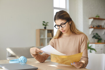 Anxious young woman at home taking letter from envelope reads bad negative news. Girl sitting at table reads notice from bank about debt on loan, about dismissal from work or notice of eviction.