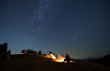 Wall Mural - Female camper resting by bonfire and enjoying pleasant atmosphere in camp outdoor. Two tents, wooden table between them and black jeep parked on hill. Beautiful shining stars in the night sky.