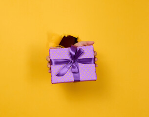 Wall Mural - female hand holds a box with a gift on a yellow background, part of the body sticks out of a torn hole in a paper background. Congratulation, holiday surprise.