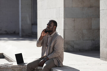 Wall Mural - Portrait of black man speaking by smartphone outdoors in city lit by sunlight, copy space