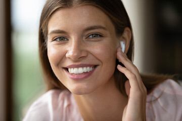 Smiling young woman with wireless earbud close up face portrait. Student girl, video talk caller, employee, operator using modern earphone, looking at camera with perfect toothy smile.