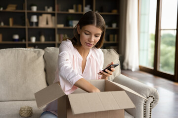 Young woman, shopper, consumer receiving parcel, unboxing package from internet store, producing goods from cardboard box, using shopping app on mobile phone, giving feedback to order delivery service