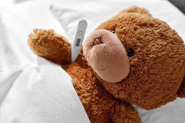 Wall Mural - medicine, healthcare and childhood concept - ill teddy bear toy head with thermometer lying in bed