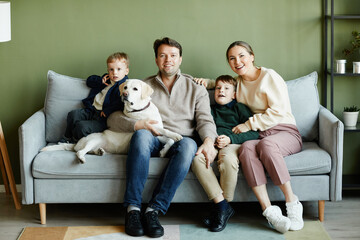 Full length portrait of big happy family with two kids and dog sitting on couch at home against green wall, copy space