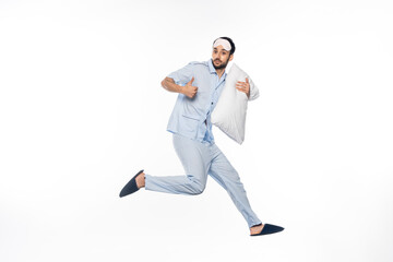 bearded man in pajamas and sleeping mask levitating with pillow while showing thumb up on white.