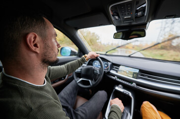 Close up view of driver behind wheel of car. Bearded young man sitting comfortably in driver's seat, placing one hand on steering wheel, the other on gearbox and staring at road ahead.