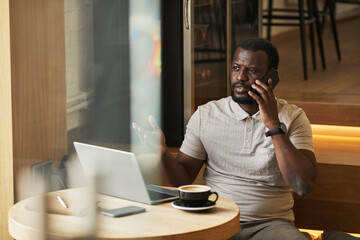 Wall Mural - Portrait of adult black man speaking by smartphone while working at cafe table in wooden interior, copy space