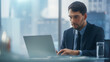 © Gorodenkoff - Confident Businessman in a Suit Sitting at a Desk in Modern Office, Using Laptop Computer, Next to Window with Big City with Skyscrapers View. Successful Finance Manager Planning Work Projects.