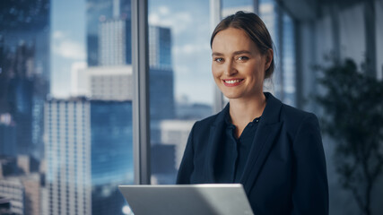 Canvas Print - Successful Businesswoman in Stylish Dress Working on Laptop, Standing Next to Window in Big City. Confident Female CEO Analyze Financial Projects. Manager at Work Planning Marketing Campaign.