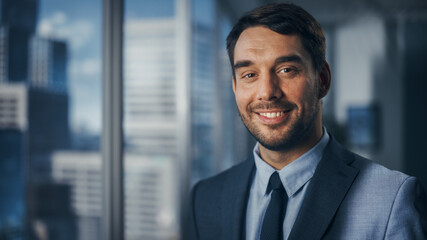 Wall Mural - Close Up Portrait of Handsome Caucasian Businessman in Stylish Black Suit Posing Next to Window in Big City Office with Skyscrapers. Confident Male CEO Smiling. Successful Masculine Business Manager.