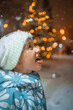 © Анна Молько - a cute child in a white fluffy hat catches snowflakes with his tongue. winter evening with garlands and snowfall.