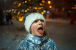 © Анна Молько - a cute child in a white fluffy hat catches snowflakes with his tongue. winter evening with garlands and snowfall.