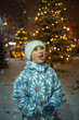 © Анна Молько - a cute child in a white fluffy hat catches snowflakes with his tongue. winter evening with garlands and snowfall.