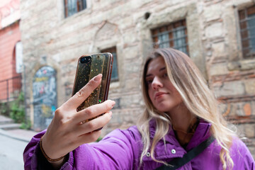 A young girl is taking a selfie with smart phone in front of a historical building. Urban life concept. City street. Selfie mania! She is wearing purple coat.