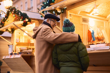 Wall Mural - Embraced grandfather and grandson buying street food at Christmas market stall.