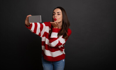 Wall Mural - Cute beautiful smiling young woman is making a selfie picture on her smartphone while posing on gray background