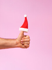 Wall Mural - Santa Claus hat is put on the finger on pastel pink background. Hand is holding thumbs up. Creative Christmas and New Year idea. Minimal announcement or like idea. Social network concept.