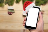 Fototapeta Sypialnia - Hand holding smartphone with a blank screen and Santa hat on Christmas background