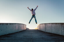Young Caucasian Man In Jeans And Hoodie Jumping With Spread Out Arms On Concrete Bridge. Mid Air Parkour Pose In City Environment And Clear Sky