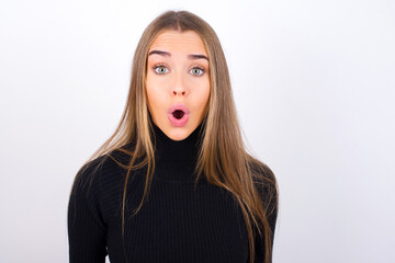 Emotional attractive Young caucasian girl wearing black turtleneck over white wall with opened mouth expresses great surprise and fright, stares at camera. Unexpected shocking news and human reaction.