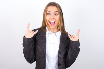 Young business woman wearing jacket over white background makes rock n roll sign looks self confident and cheerful enjoys cool music at party. Body language concept.