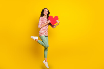 Poster - Photo of funny cute mature lady dressed striped shirt jumping high holding big red heart smiling isolated yellow color background