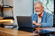 © chika_milan - The happy senior student is sitting at home and typing homework on the laptop. Retraining at old age concept.