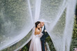 A young, stylish groom and a beautiful brunette bride embrace under a long fabric veil. Wedding portrait of newlyweds in love.