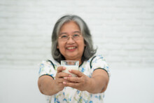 Asian Senior Woman Holding Glass Milk While Relaxing On A Sofa Living Room For Retirement Wellness. Elderly Woman Drinking A Glass Of Milk To Maintain Her Wellbeing. Concept Of Wellness And Healthy.