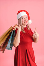 An Adult Woman In A Santa Hat With Shopping Bags Shows Her Thumbs Up Posing On A Pink Background