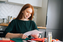 Side View Of Happy Young Woman Cutting Out Craft Paper For Envelopes With Scissors Making Christmas Advent Calendar On Winter Holidays Eve, Selective Focus, Blurred Background.