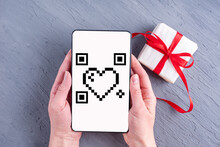 Hands Holding Mobile Phone With White Screen With Heart Shaped Qr Code And Gift Box With Red Ribbon On Gray Background. Online Shopping For Valentine's Day. Online Shopping QR Code Payment
