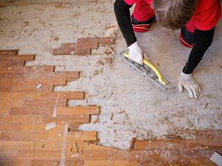 Craftsman in workwear who, using the rabot, removes excess glue left on the floor after removing the wooden parquet glued to the slab of an apartment. Worker at work while performing home renovation.