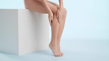 Horizontal shot of slim young attractive woman strokes her smooth leg sitting on the white platform on pale blue background | Moisturizer commercial