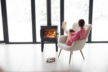 Woman sitting with cup on chair by the fireplace at modern house on nature during winter time. Concept of winter mood and comfort at home. Girl wearing hat and pink sweater